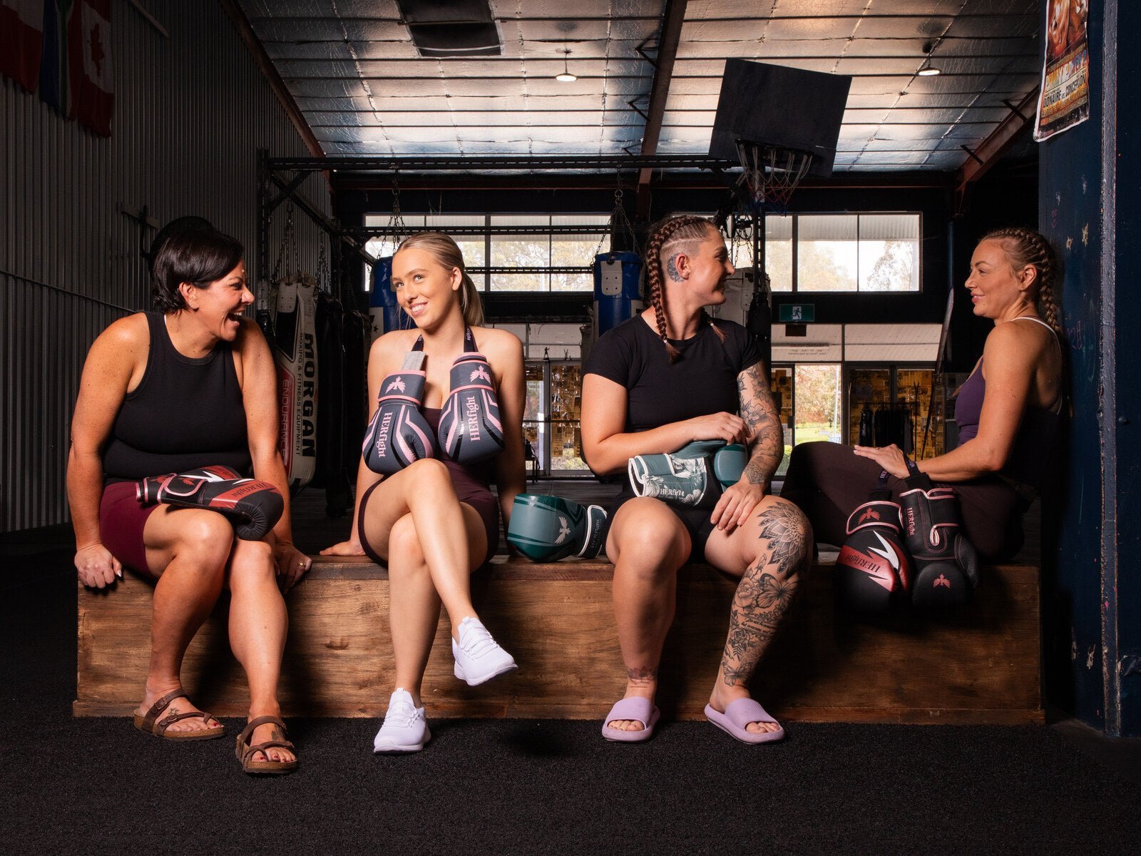 Four women of different ages and body types sitting together on a bench in a boxing gym, each holding HERfight women's boxing gloves in pink, red and teal colourways